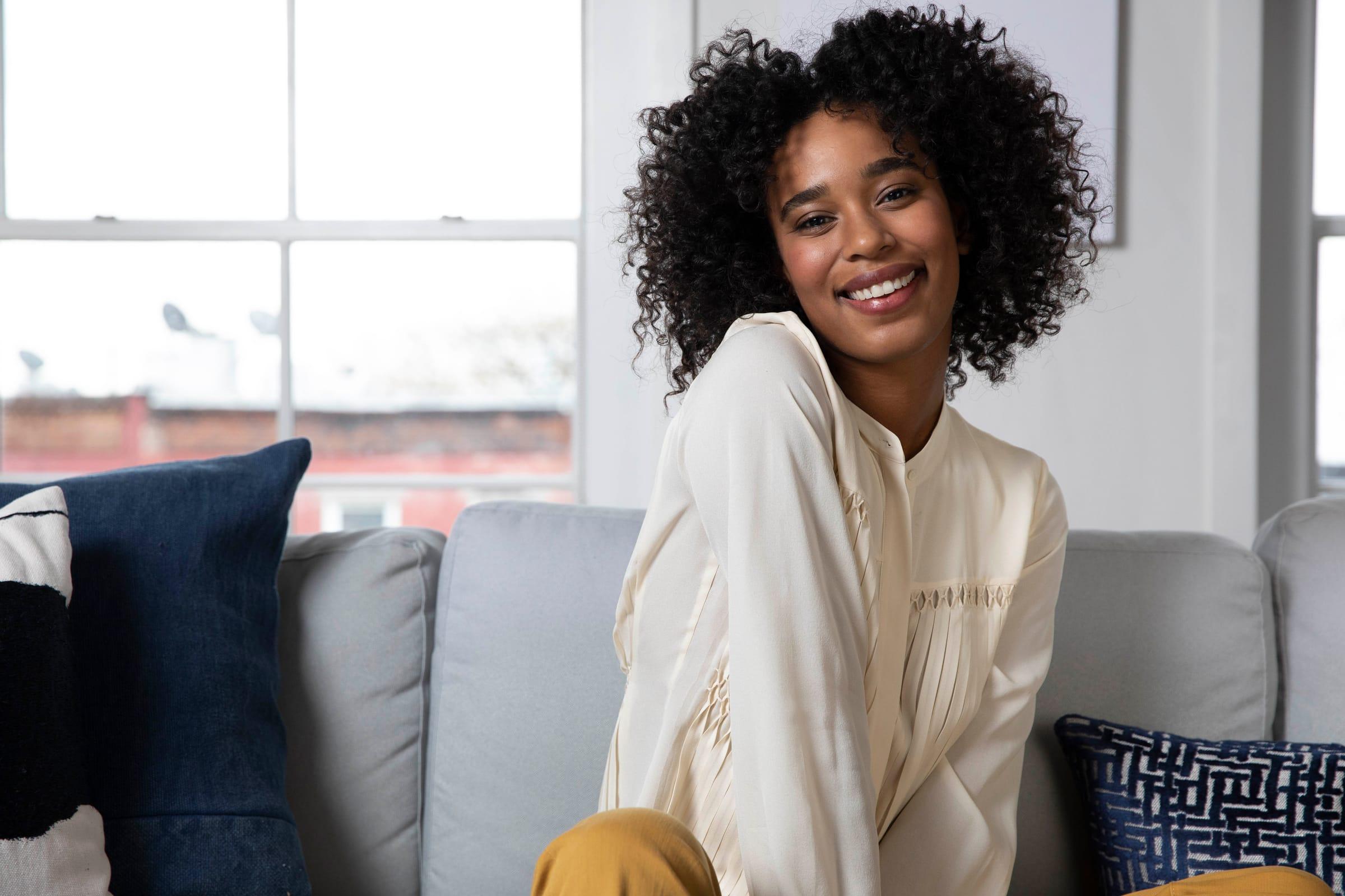 A smiling woman with curly hair seated on a sofa with navy cushions